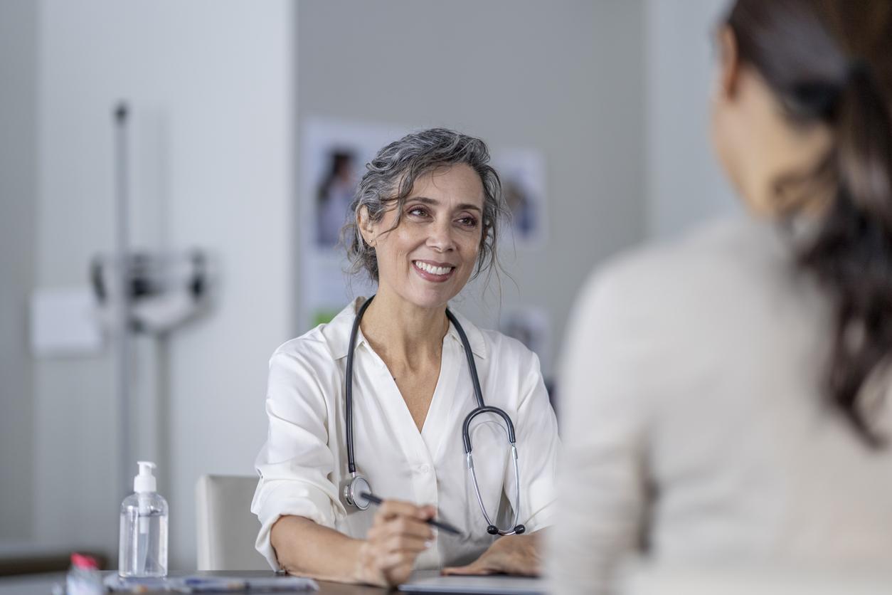 A mature female doctor points to the screen of her tablet laying out on the table in front of her as she shares her patients test results with her.  She is wearing a white lab coat and has a stethoscope around her neck as she talks with the female patient of Asian decent.